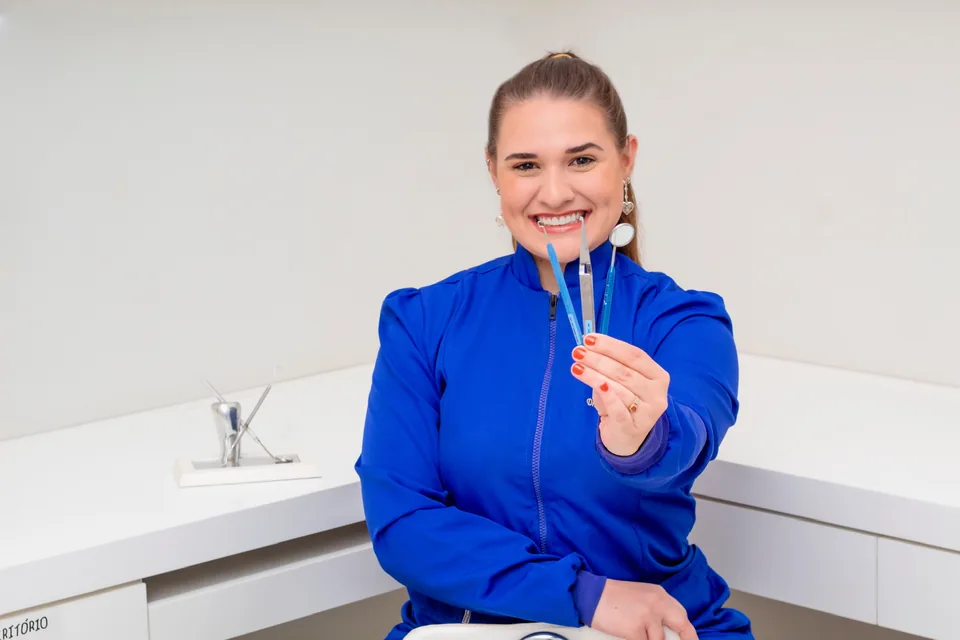 Dra. Lara Gimenez de uniforme azul, sorrindo e segurando instrumentos odontológicos no consultório