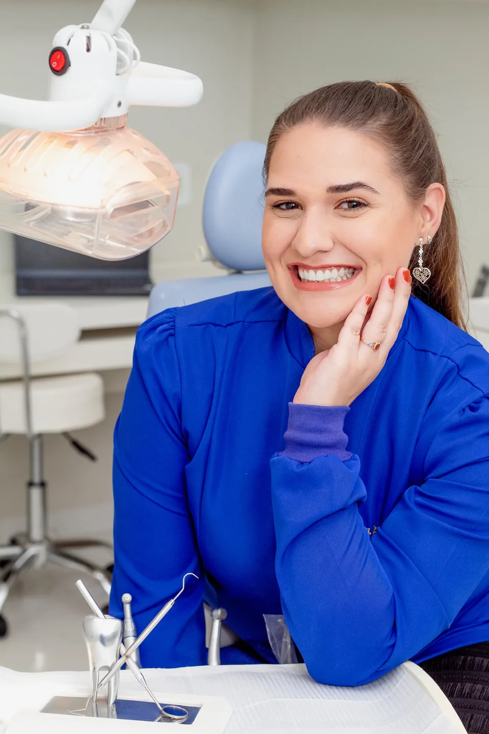 Dra. Lara Gimenez de uniforme azul, sorrindo ao lado da cadeira odontológica no consultório